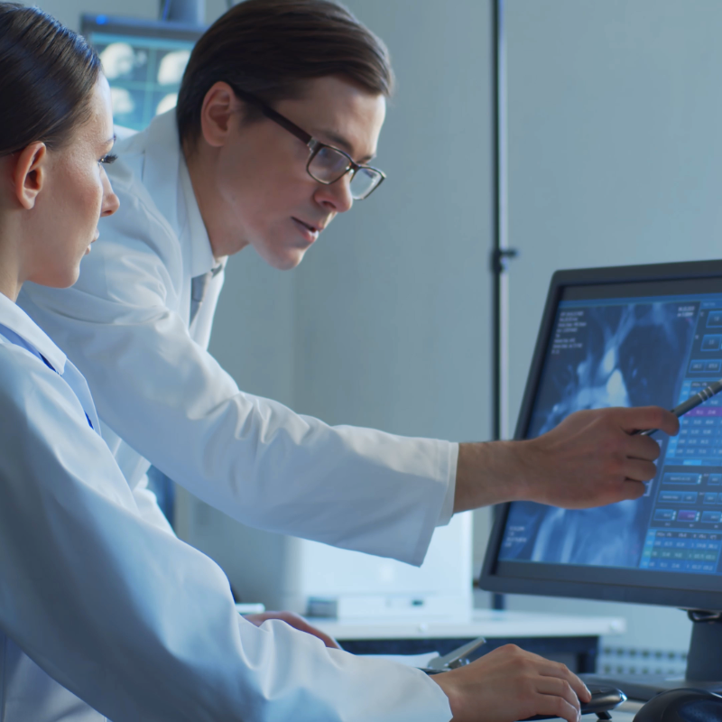 Two people in lab coats review medical imaging on a computer monitor, with one person pointing at the screen while the other looks on.
